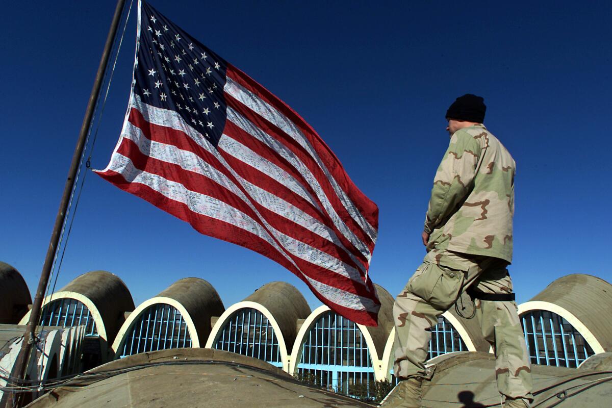 A man in camouflage stands alongside a U.S. flag.