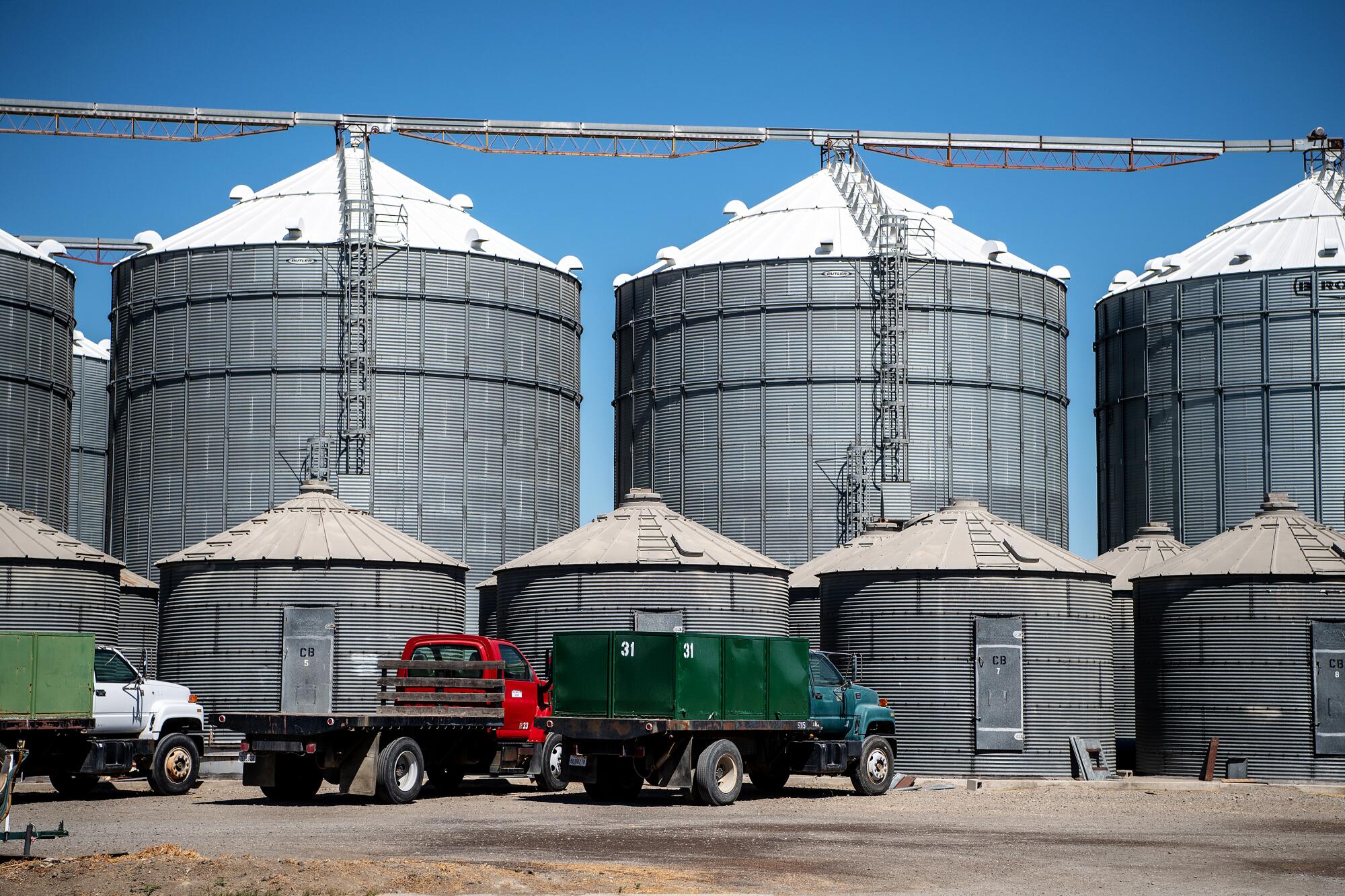 Three farm vehicles are parked next to grain silos