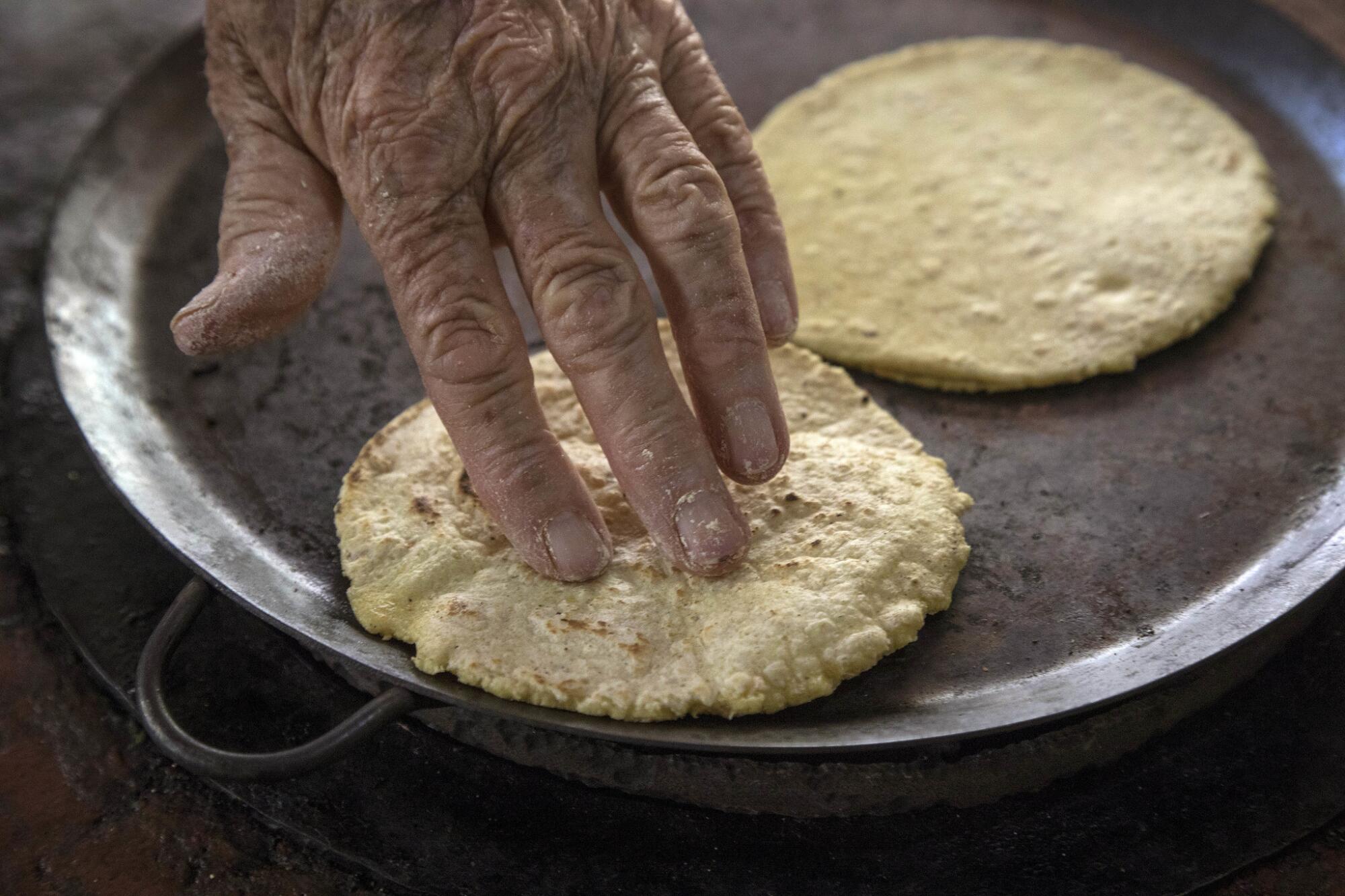 A wrinkled hand presses a tortilla on a comal.
