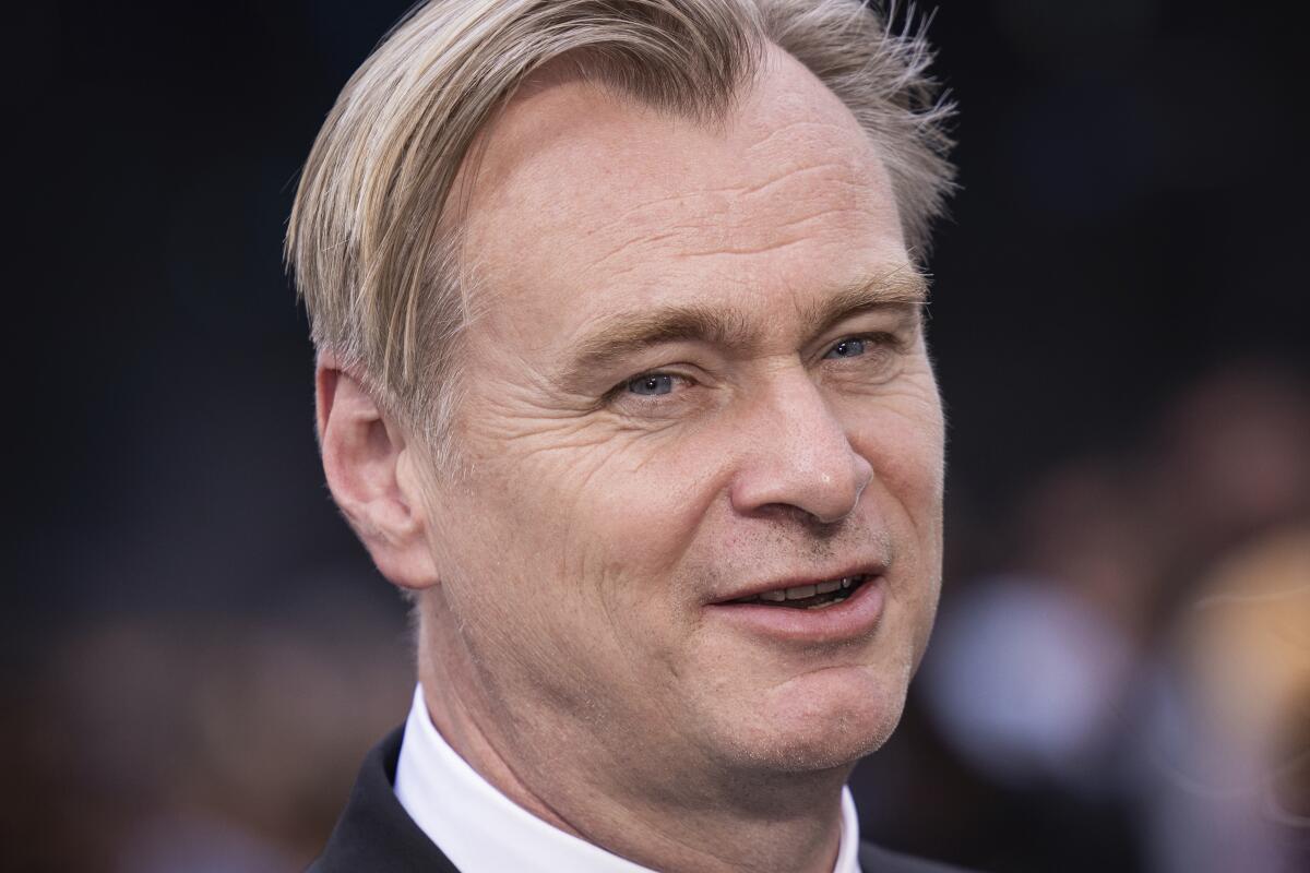 Christopher Nolan smiles and looks off to the side in a black suit with white shirt collar