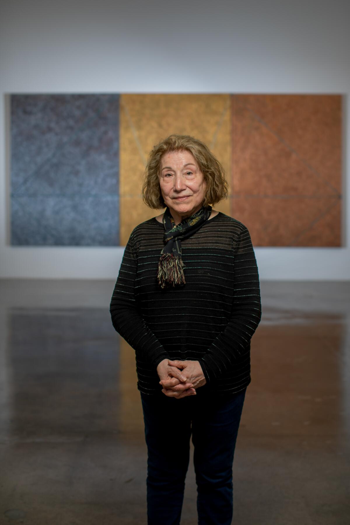 Virginia Jaramillo, dressed in black with scarf tied around her neck, stands before a tricolor abstract painting. 