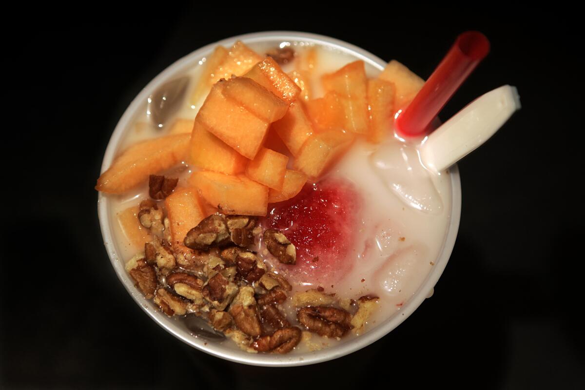 A plastic cup with a straw and spoon holds a drink with sliced melon and nuts.