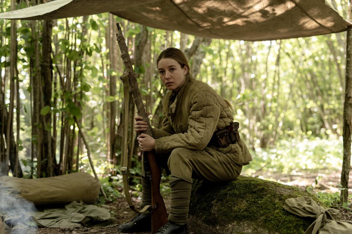 A woman in World War II military gear sits on a log holding a rifle in the movie "Burial."