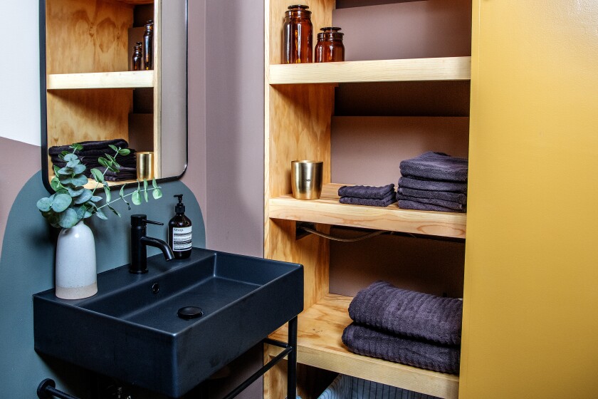 A sing and mirror are mounted on a purple and gray-blue wall, kitty corner to wooden shelving with towels on it.