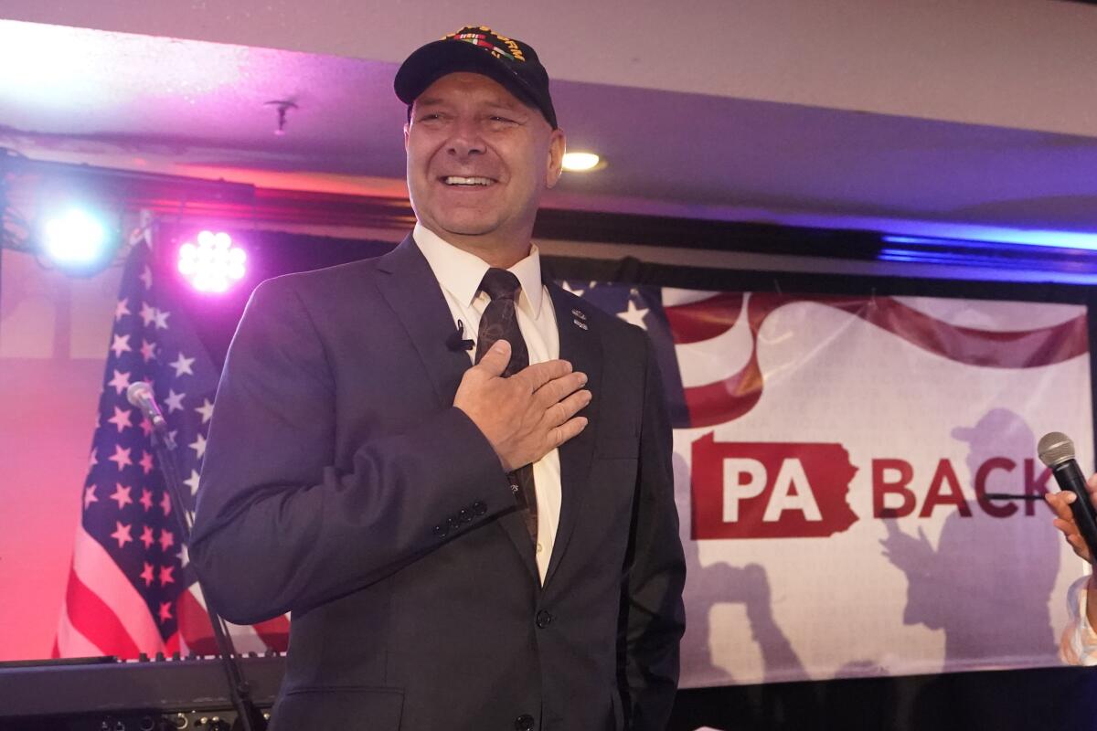 State Sen. Doug Mastriano puts his hand on his chest and smiles in front of a U.S. flag and lights.