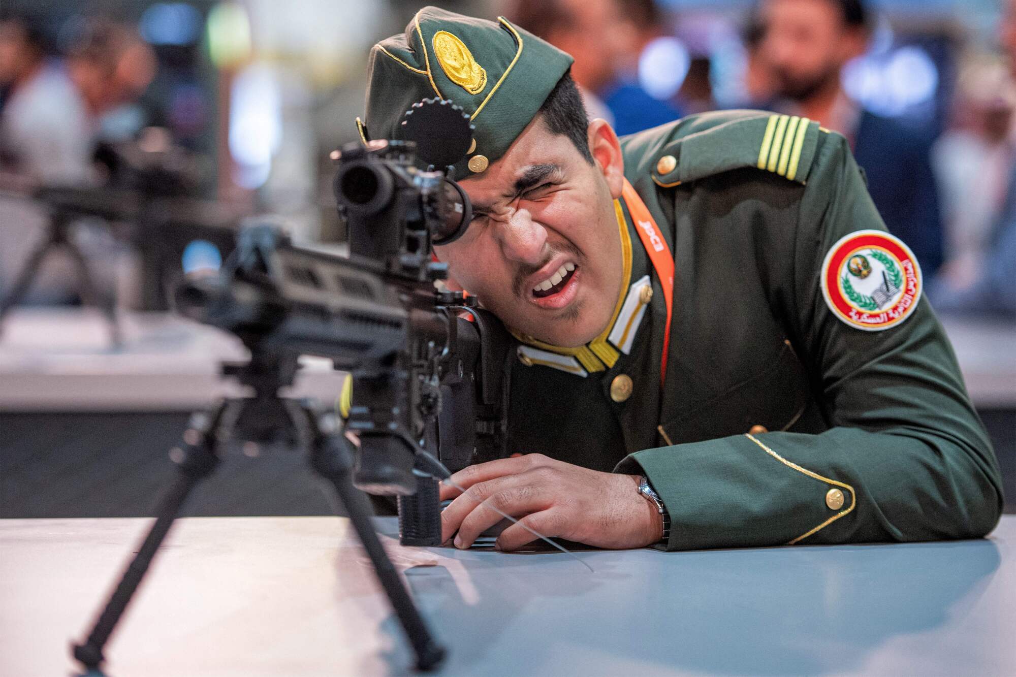 Un homme en uniforme militaire vert regarde à travers la portée d'un fusil d'assaut exposé 