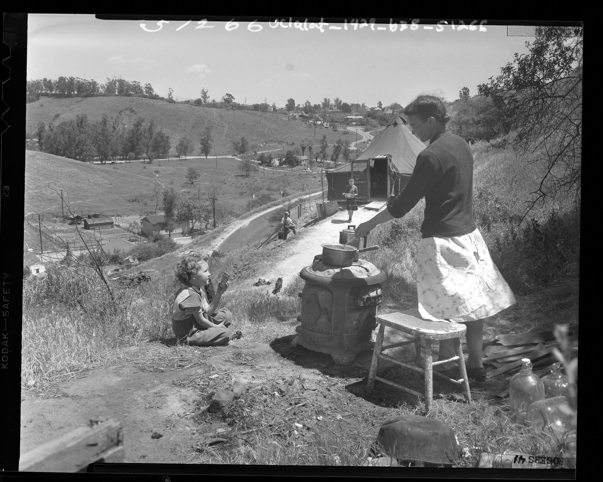 A woman prepares a meal on a cookstove as one child plays and others struggle up a hill while carrying water in 1948.