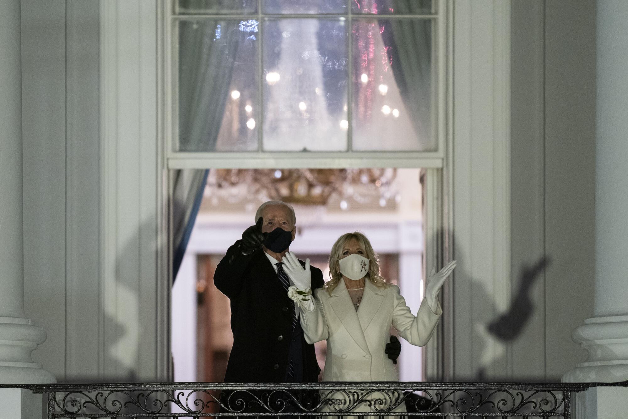 On a White House balcony, Jill Biden holds both gloved hands open as Joe Biden points toward the sky.