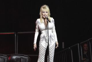 Dolly Parton singing into a mic wearing white pants suit with silver chains. 