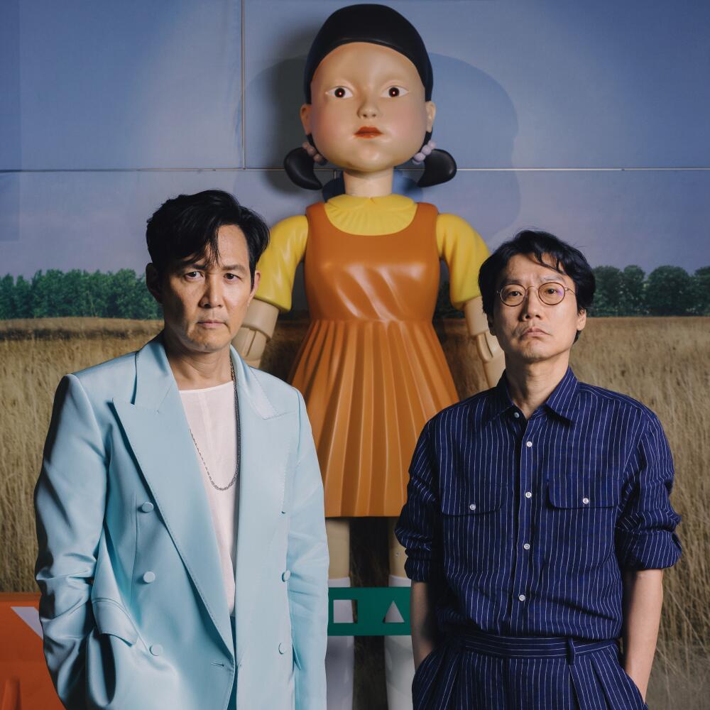 A man in a light blue suit stands next to a man in a blue pinstripe shirt. A doll in an orange dress towers behind them.