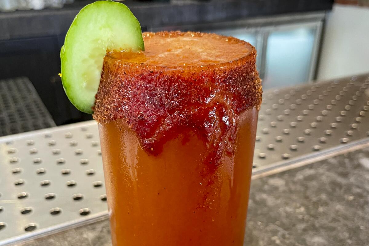A glass containing a red-hued michelada, with a red salty rim and a slice of cucumber for garnish.