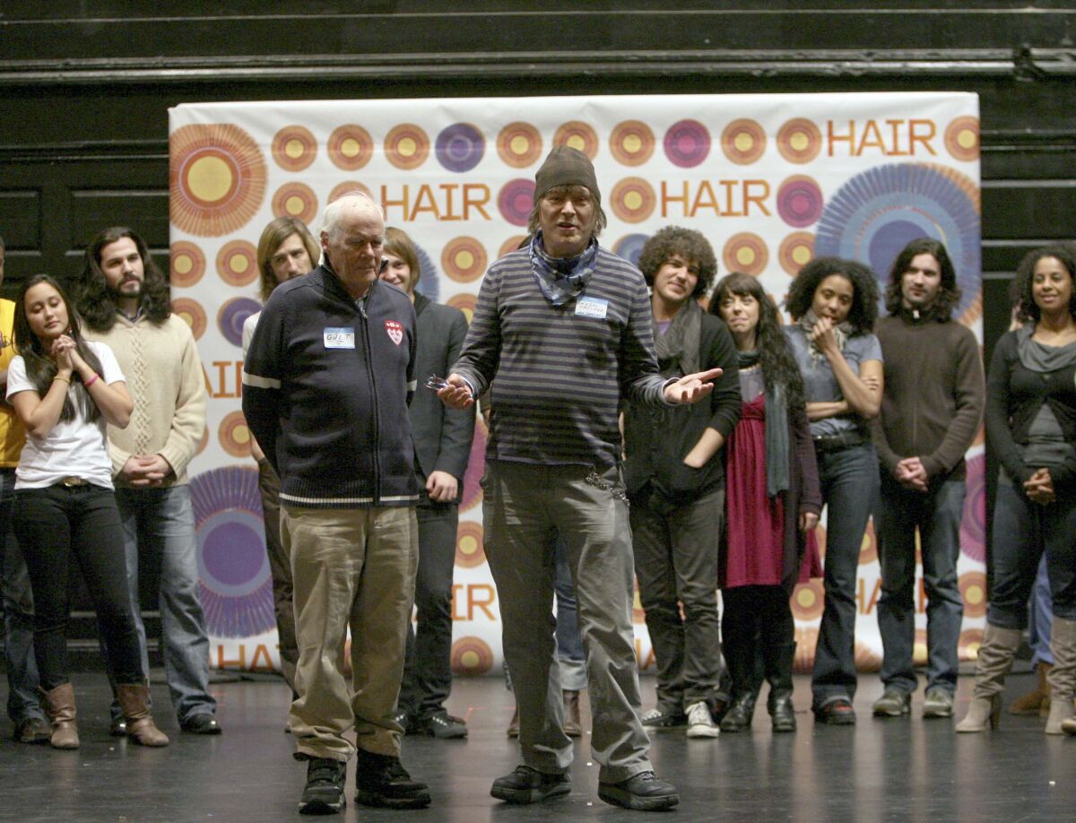 Two older men stand in front of a troupe of younger performers onstage