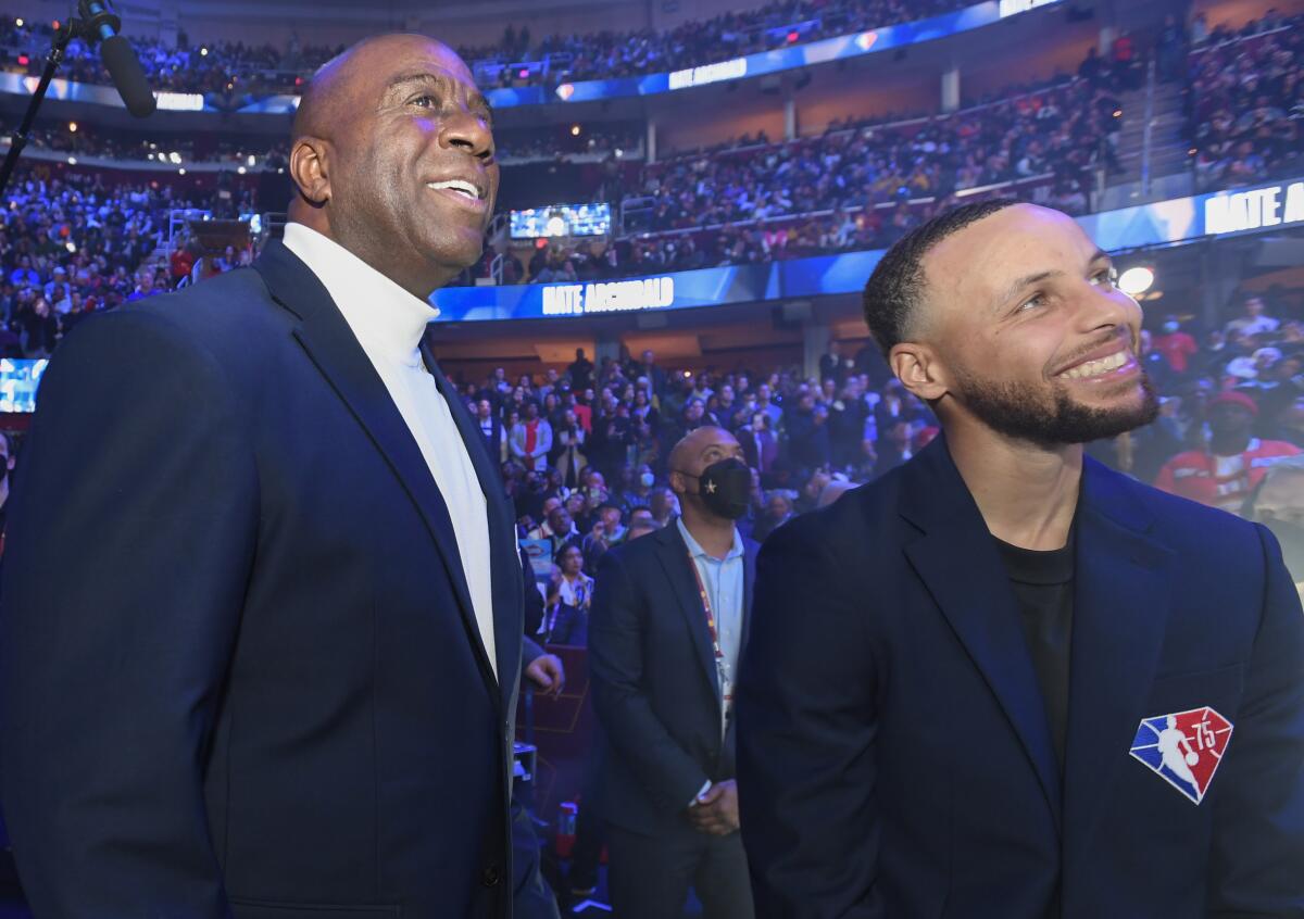 Magic Johnson and Stephen Curry smile in an arena filled with people.