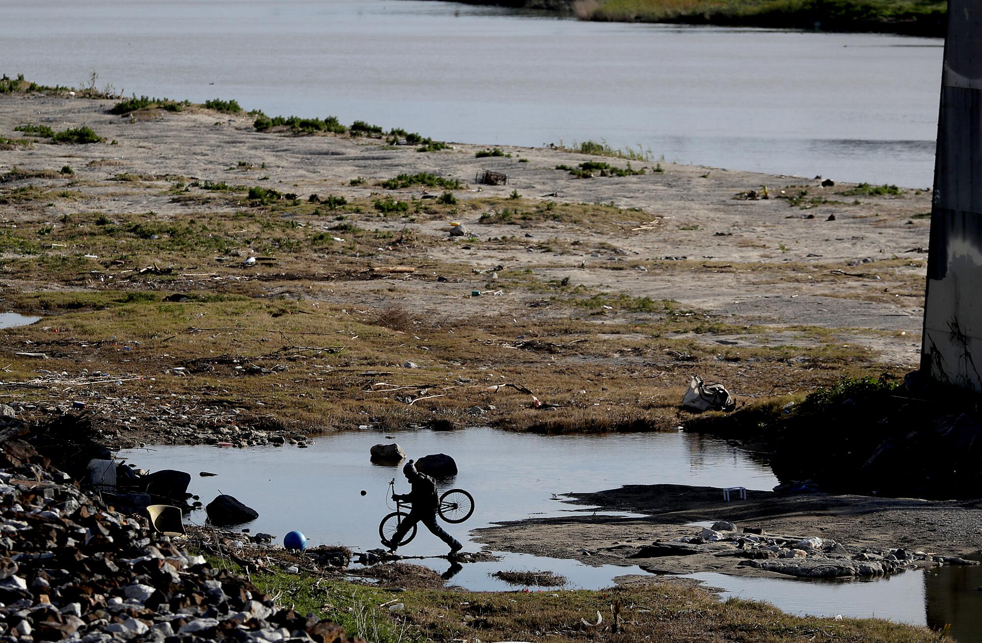 A man, holding a bicycle, hops over a puddle near the Los Angeles River