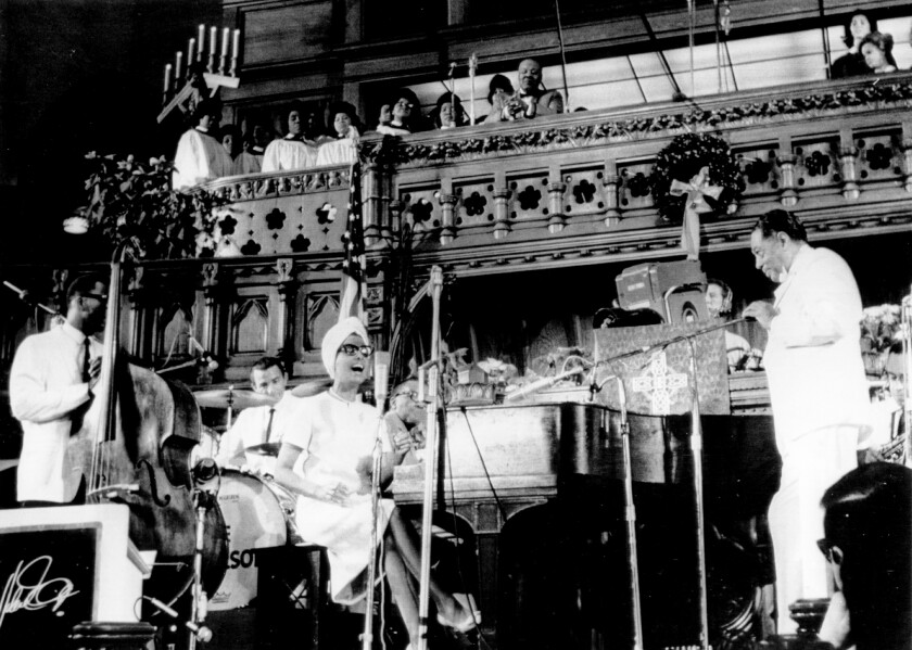 A seated woman sings as a pianist, bassist and drummer perform; a choir is seen on a balcony above them.