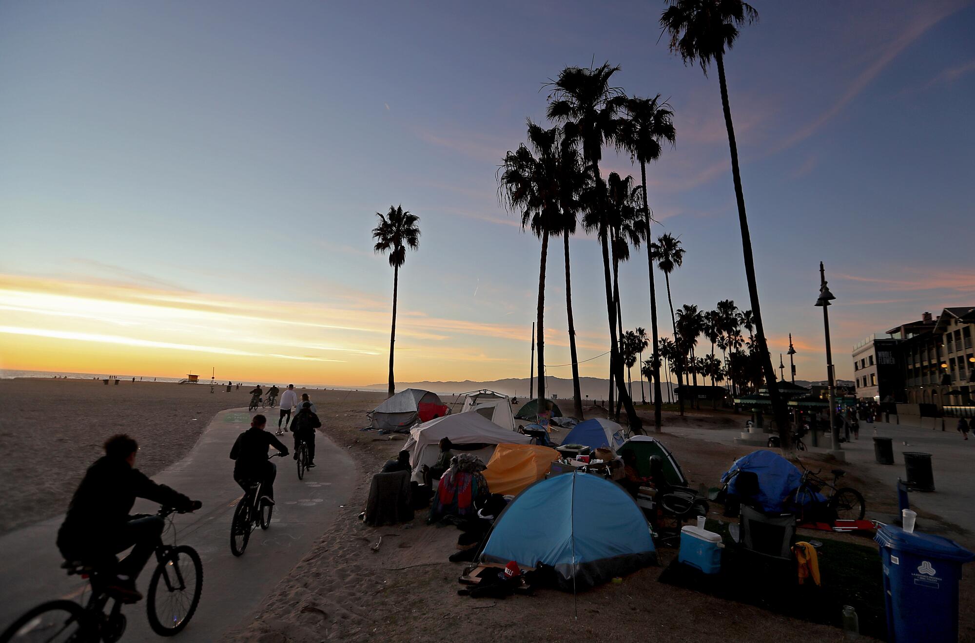 People ride bike at sunset as they pass a homeless encampment in Venice Beach.