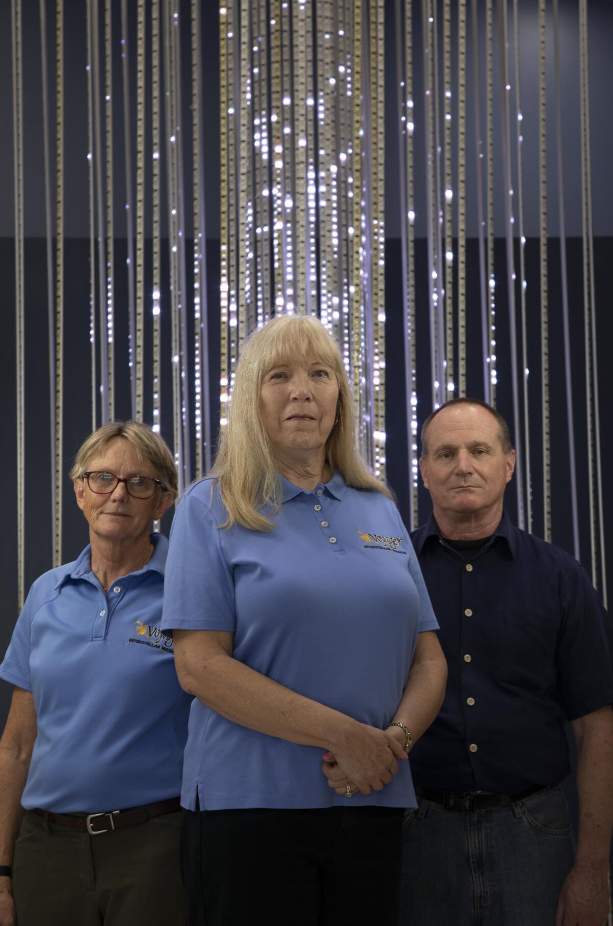 Two women in light blue shirts stand next to a man in a dark shirt 