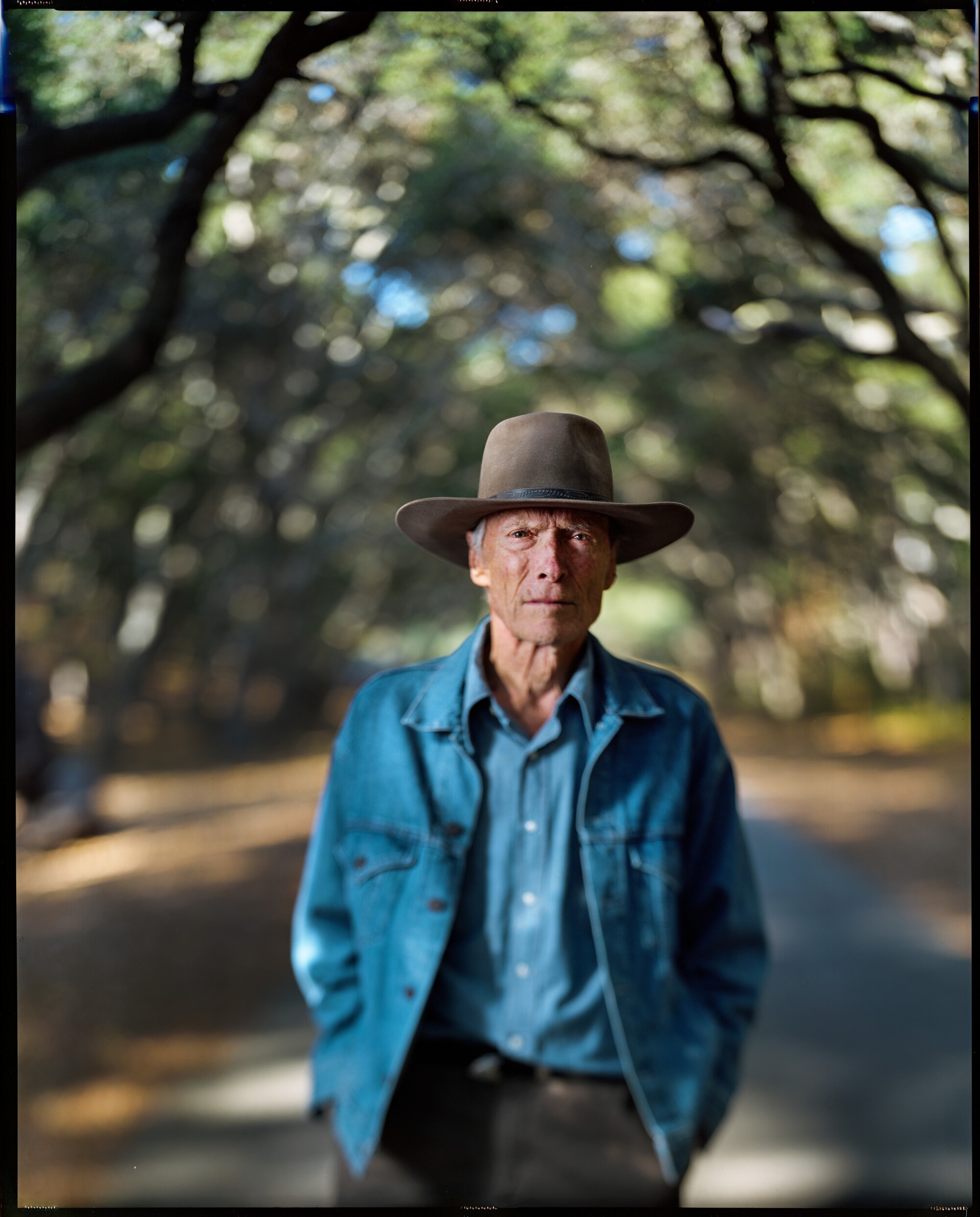 A man in a denim jacket and brimmed hat stands under oak trees for a portrait.