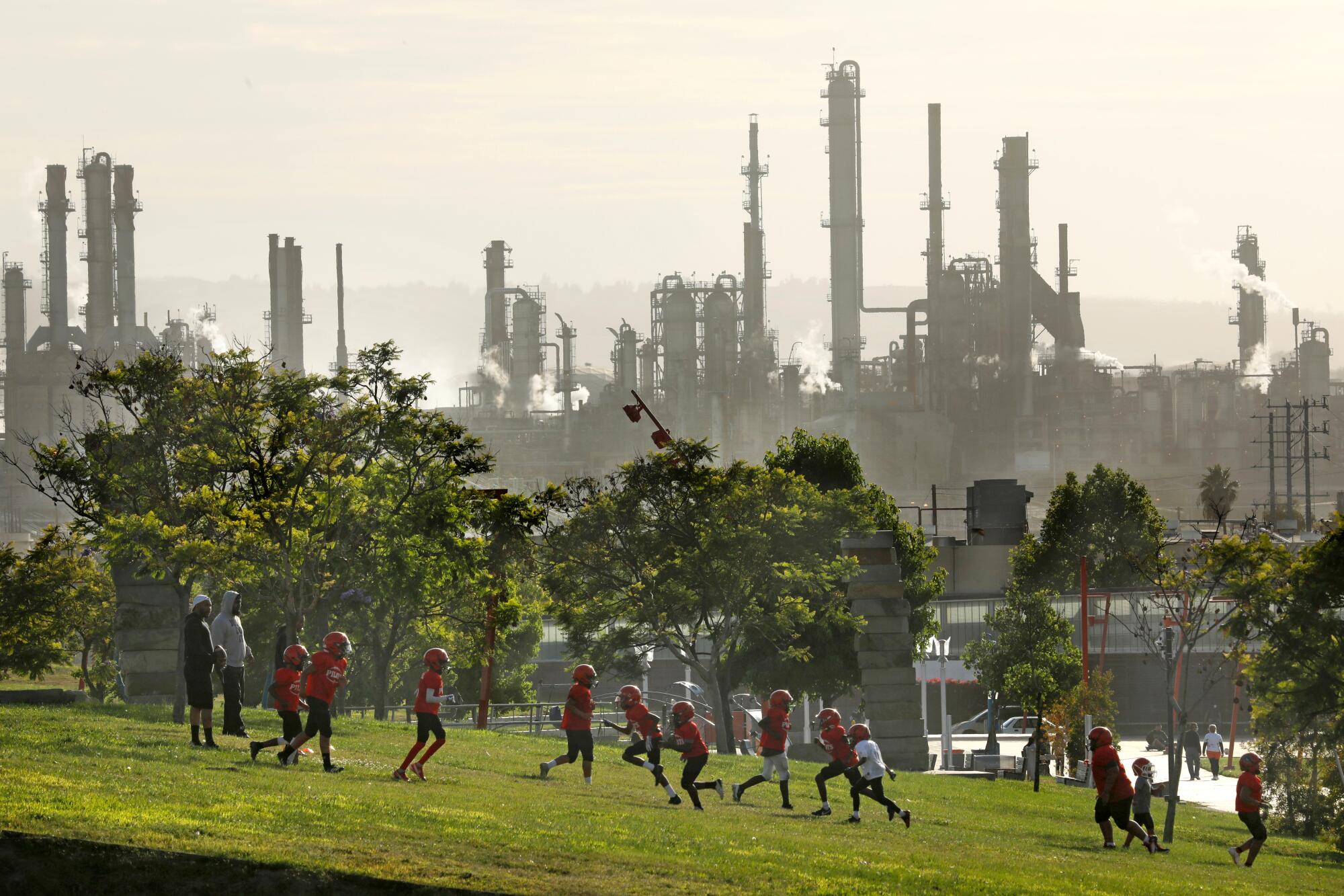 People in red helmets and sports uniform run in a grassy area, with a refinery as a backdrop