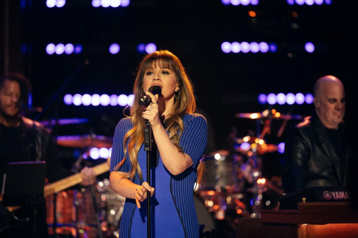 Kelly Clarkson with long wavy hair and bangs singing into a microphone while clad in a blue short-sleeve dress 