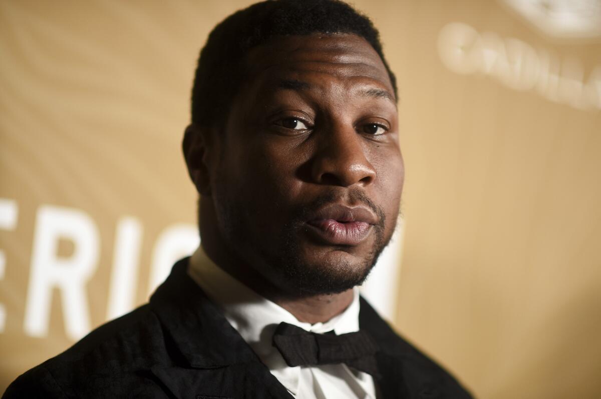 Jonathan Majors looks forward with pursed lips in a tuxedo against a gold background