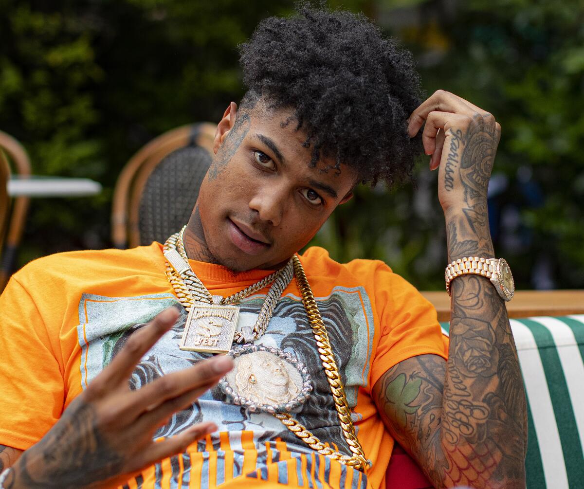 Blueface, in an orange T-shirt and gold chains, throws a sign with one tattooed hand while touching his hair with the other