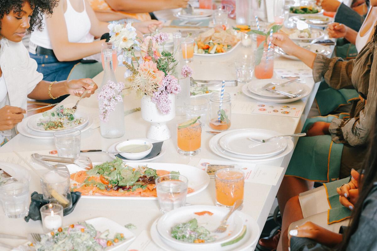 A group of women chat over a meal. Flowers in vases and plates of food cover the table.