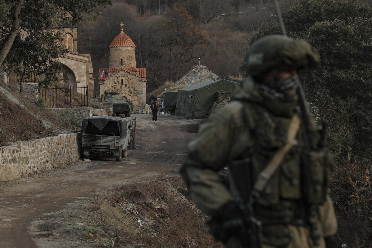 A man in full military gear, carrying a rifle, stands near a monastery. In the background are military vehicles and tents. 