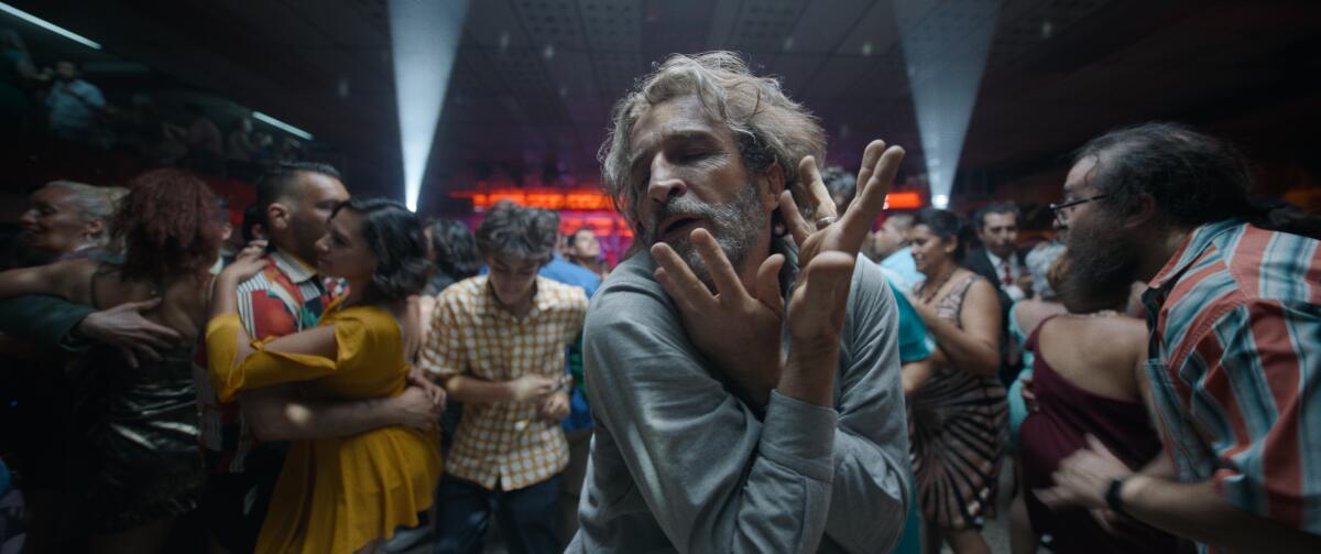 A man holds his hands up to his face as people dance around him in the movie "Bardo"