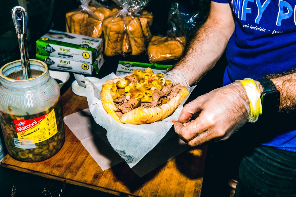 A pair of hands holds an Italian beef sandwich piled with giardiniera amid a sandwich-making station.