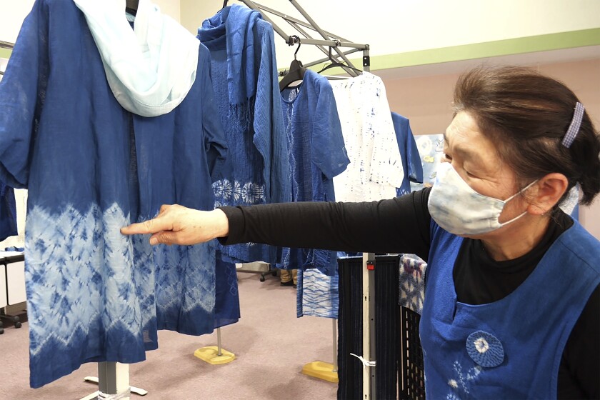 In this image from video, Kiyoko Mori, 65, the head of indigo dye group called Japan Blue, points out at one of displayed indigo dyed artwork at a community center where residents evacuated when the 2011 earthquake hit the area in Minamisoma, Fukushima Prefecture, northeastern Japan, on Feb. 20, 2021. After the Fukushima nuclear plant disaster a decade ago, nearby farmers weren't allowed to grow crops for two years because of radiation. After the restriction was lifted, two farmers in the town of Minamisoma found an unusual way to rebuild their lives and help their destroyed community. Kiyoko Mori and Yoshiko Ogura planted indigo and soon began dying fabric with dye produced from the plants. (AP Photo/Chisato Tanaka)