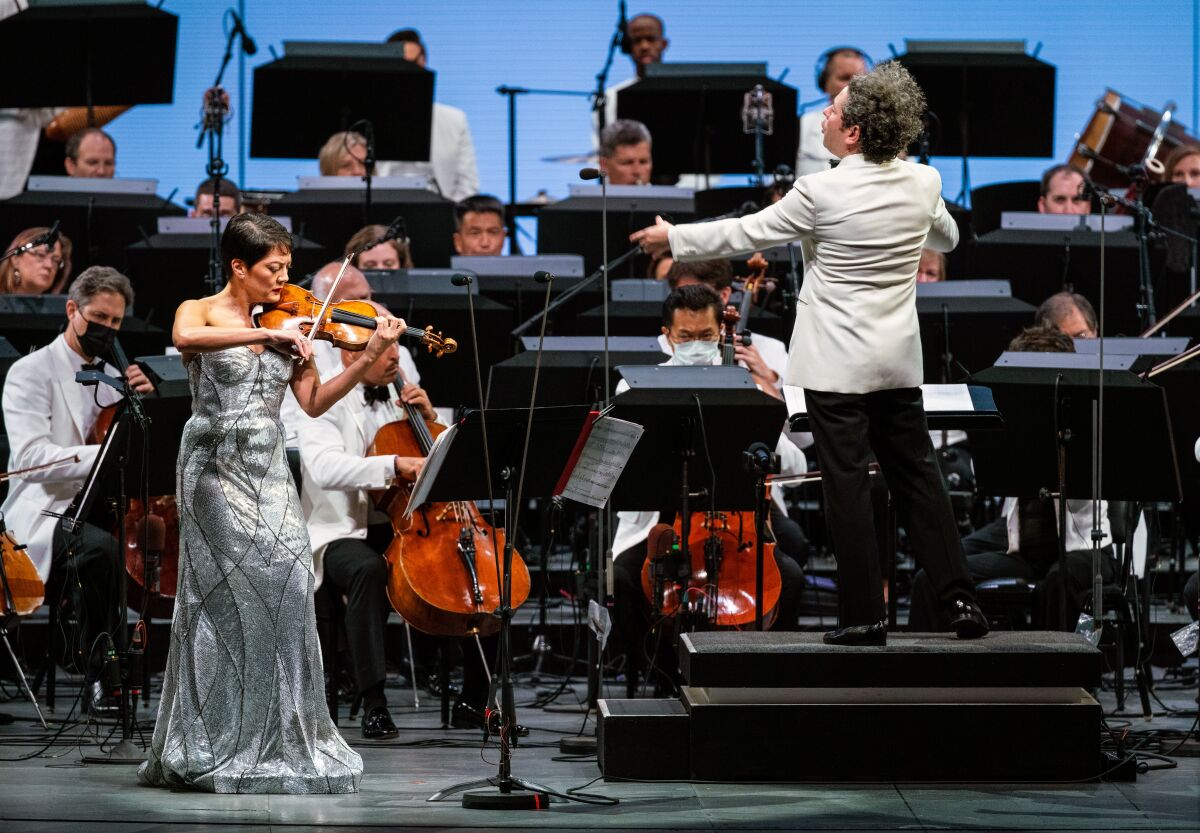 A woman in a silver-colored dress plays violin as a man conducts and orchestra