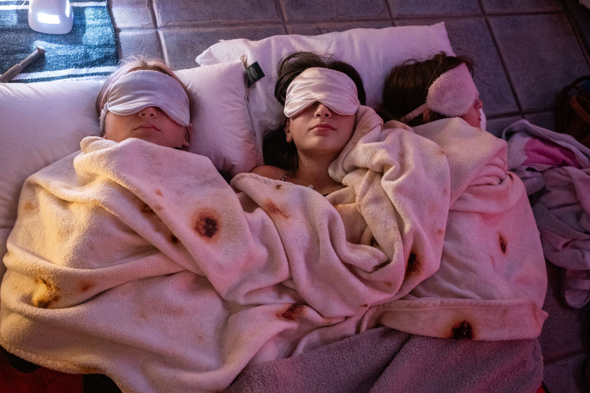 Three people wearing face masks and sharing a blanket and pillows relax during a sound bath.