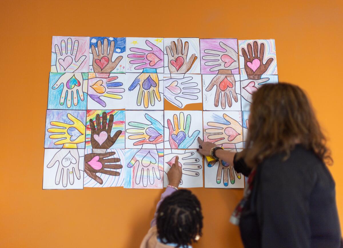 A teacher and a young student point at a collage of children's artwork depicting hands with hearts in them.