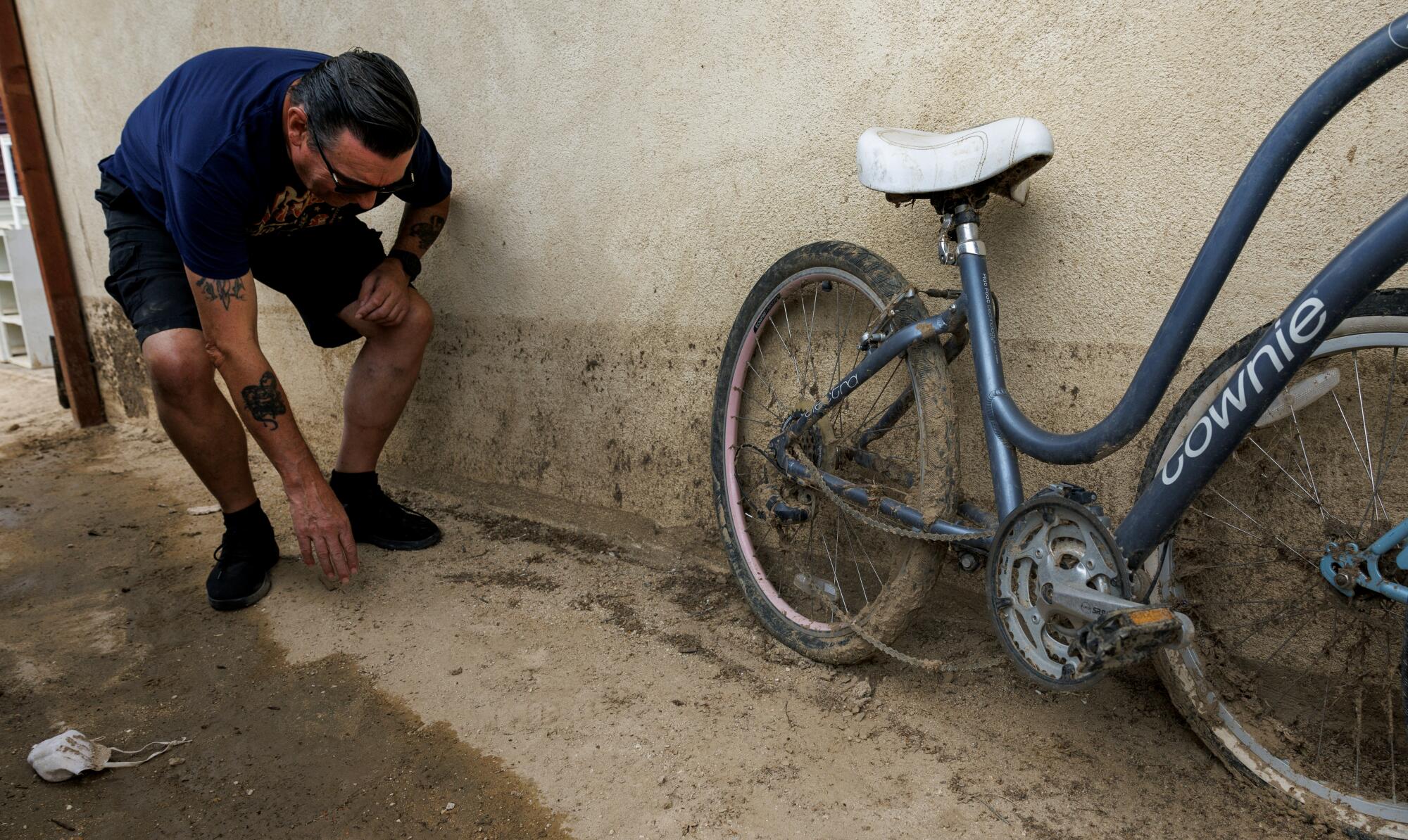 A man with dark hair, in dark clothing, bends down with one hand touching the ground, near a muddy bicycle against a wall