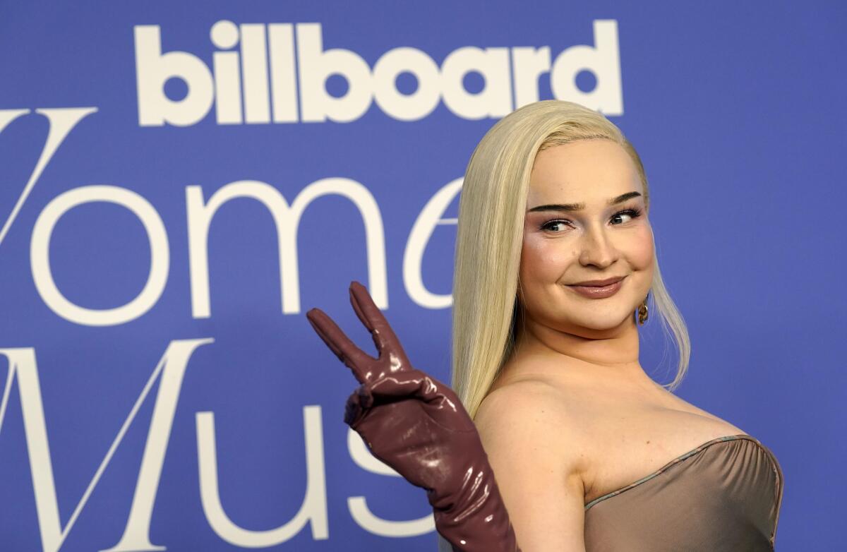 Kim Petras in a copper strapless dress holding up a peace sign with her right hand, which is in a brown patent leather glove