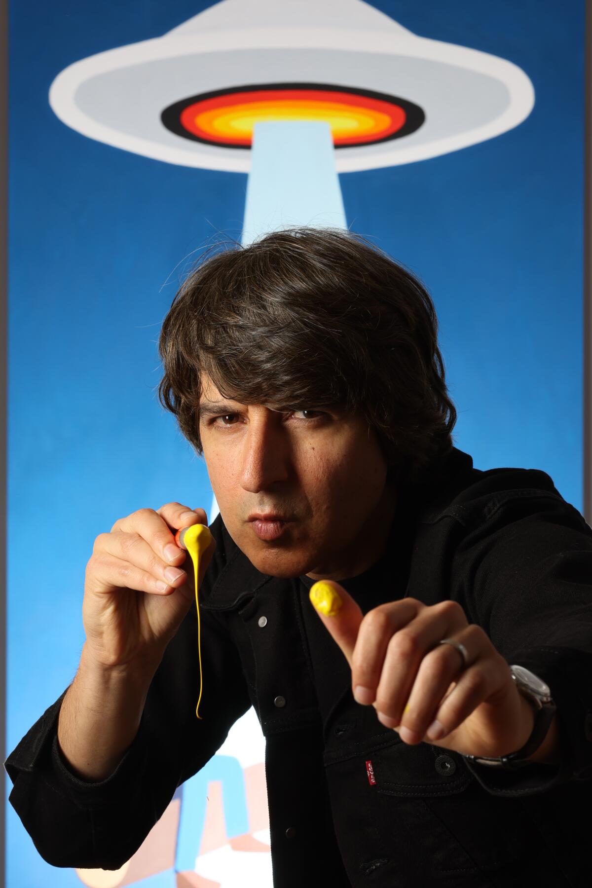 Demetri Martin stands in front of a painting of a UFO beaming up a man.