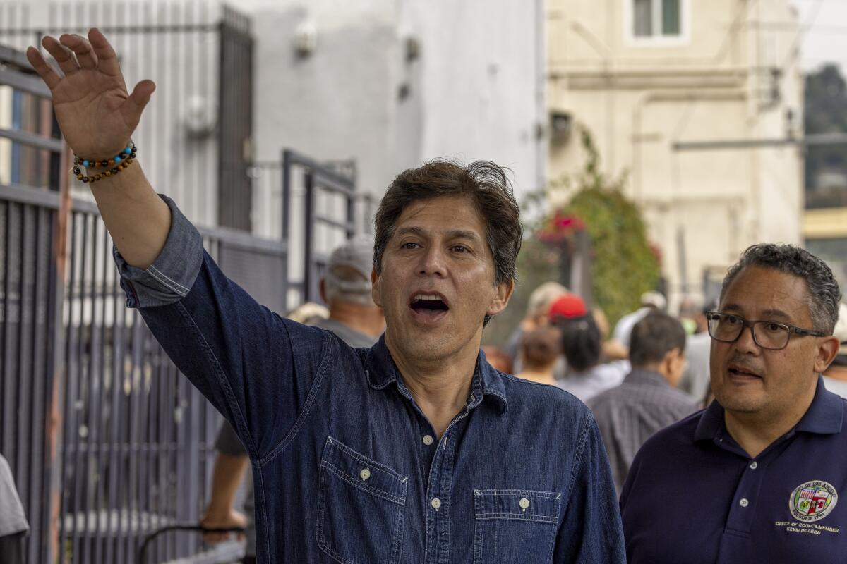 Kevin de León, in a dark-blue denim shirt, stands near urban buildings with another man and waves to someone in the distance