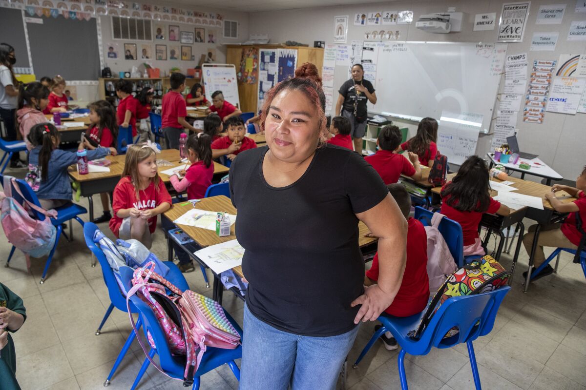 A woman stands in front of a desk in a classroom filled with children