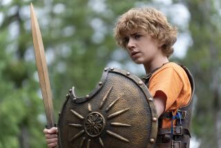 A boy with curly blond hair holding a gold shield and sword in the woods.