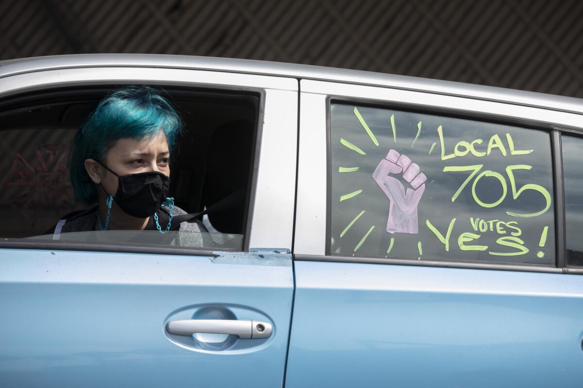  A woman wearing a face mask in the drivers seat of a car. The backseat window says "Local 705 votes yes!"