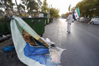 A man in white clothes holds a green, white and black flag on a street, near a large poster of a man discarded at a dumpster