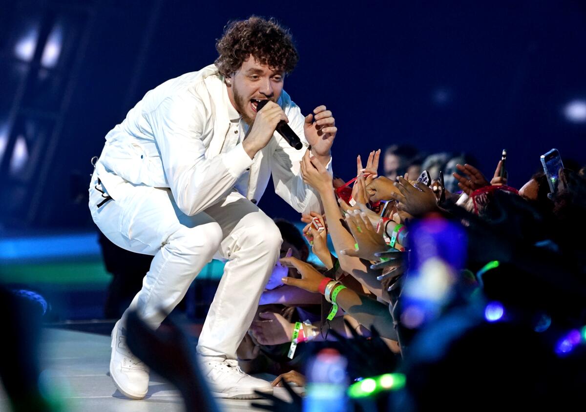 A man wearing all white and holding a microphone to his mouth crouches onstage as a sea of hands reaches toward him.