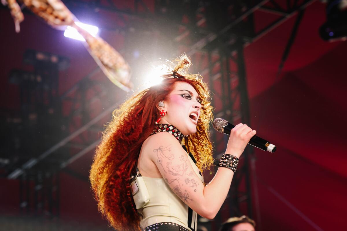 A person with long red hair sings into a microphone under lights.
