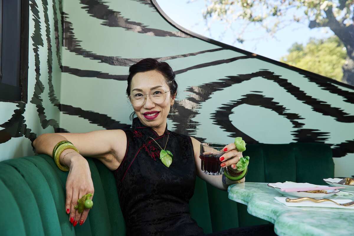 A woman sitting in a restaurant booth, holding a drink and wearing what looks like jade jewelry.