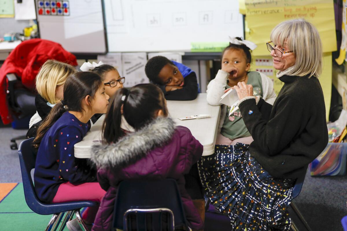 A woman wearing glasses sits at a classroom table talking with six children, holding a book.