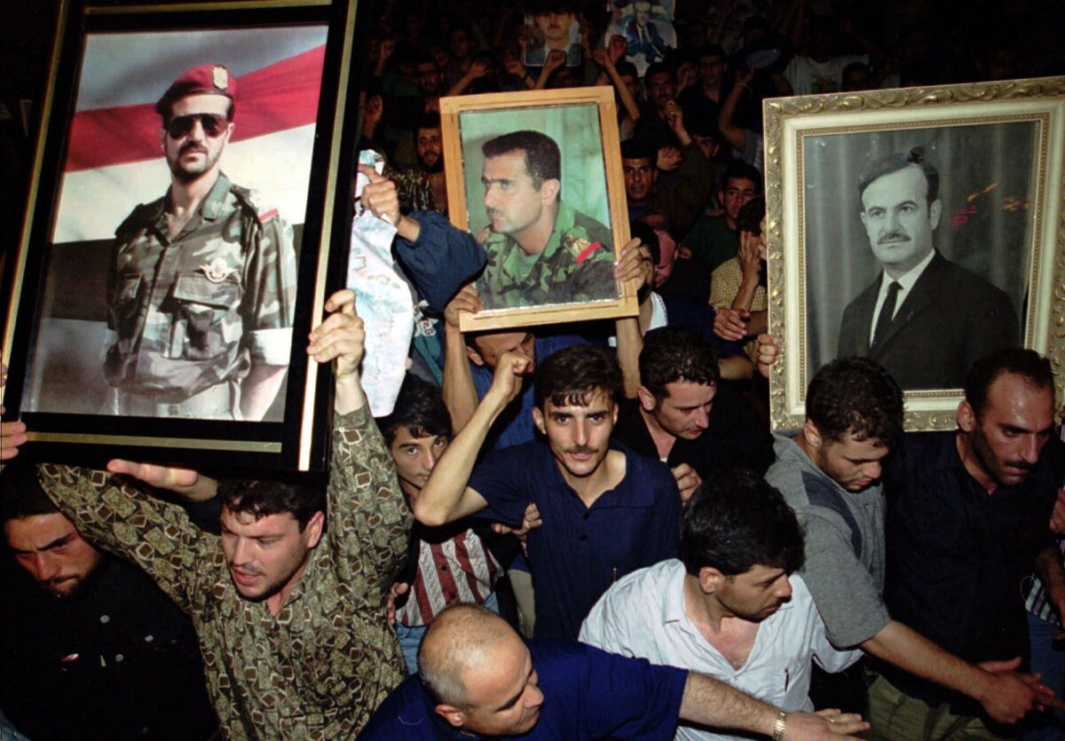 A crowd of Syrian mourners, with three of them holding up framed portraits of Assad family members