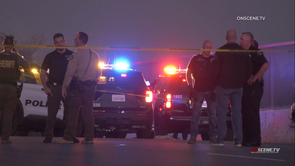 Officers stand near patrol cars with their lights on and yellow police tape blocking a street