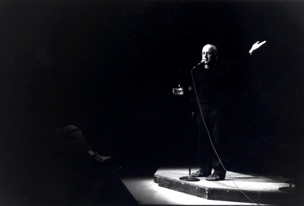 A man stands on a stage in a spotlight, gesturing with one arm raised