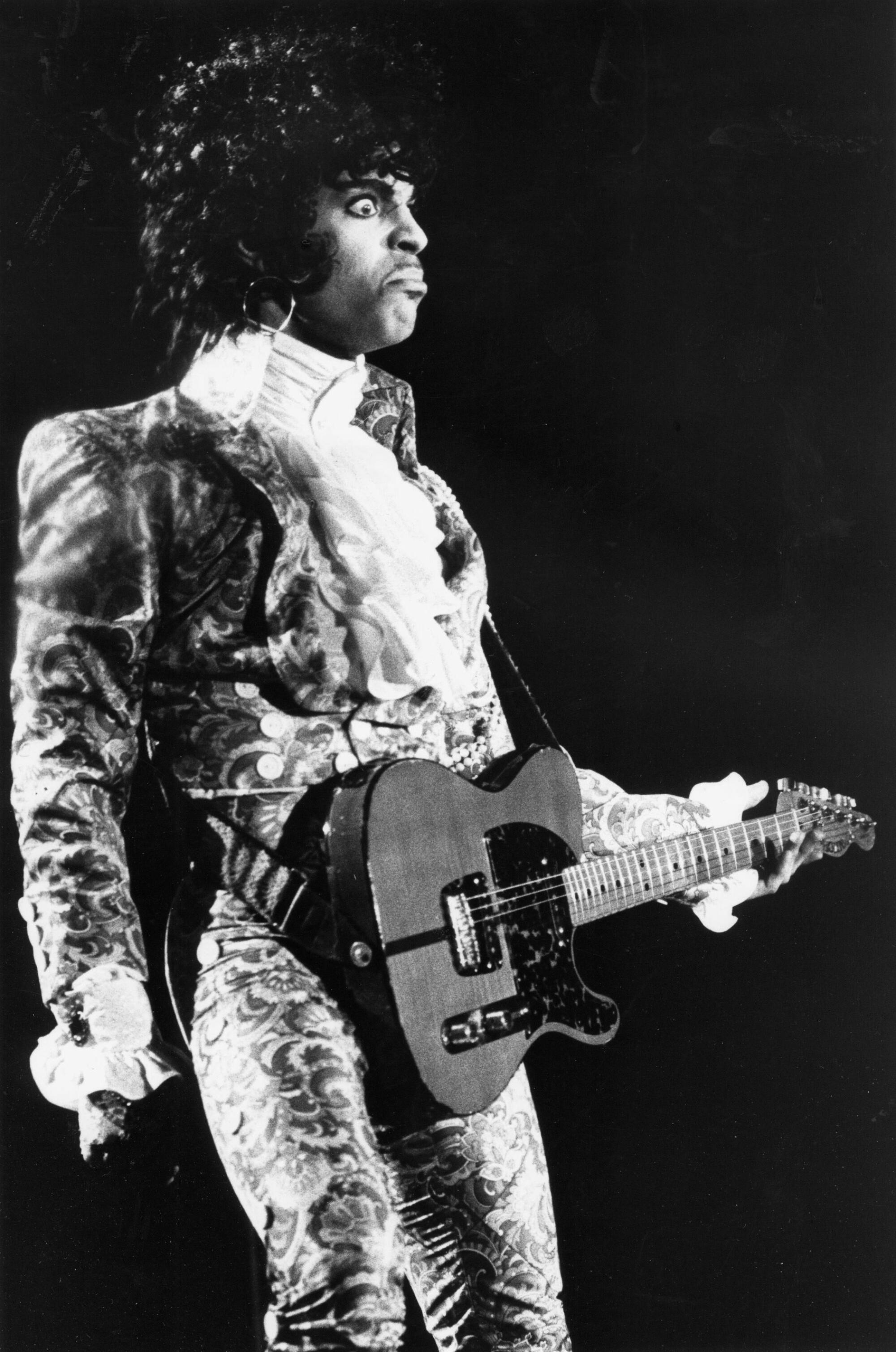 Prince, in ruffled shirt and velvet suit, holds a guitar onstage.